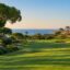 Autumn golf in the Algarve: a sunlit fairway edged with pines and palms, a flag on the green and the blue Atlantic beyond.