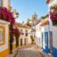 Lagos old town cobbled street with whitewashed houses, bougainvillea-draped balconies and a church bell tower against blue sky